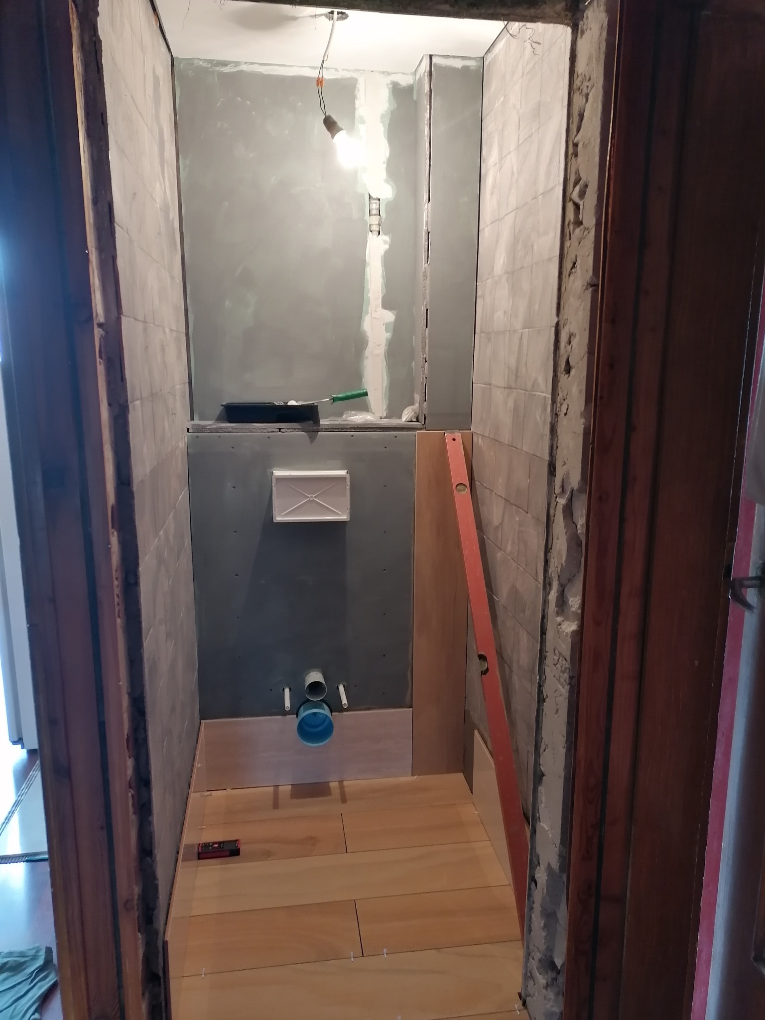 Wnętrze małej łazienki w trakcie prac w Mucharzu: częściowo położone płytki na ścianach, podłoga wyłożona panelami, widoczne instalacje i poziomica.