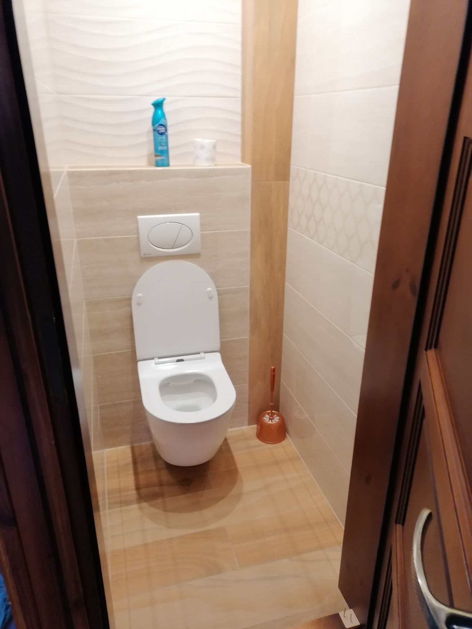 Nowoczesna toaleta w beżach z podwieszaną muszlą, przyciskiem spłukującym w zabudowie, ceramicznymi płytkami imitującymi drewno na podłodze i ścianach, oraz akcesoriami łazienkowymi w postaci...