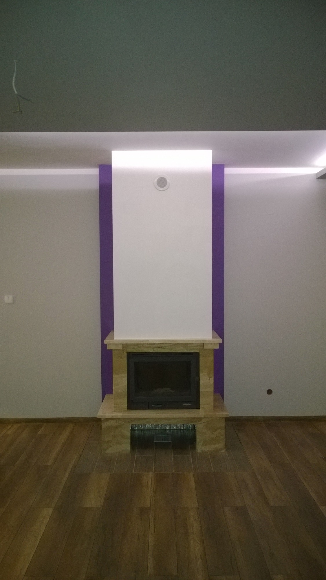 Wykończony kominek z obudową z jasnego marmuru, otoczony fioletowymi pasami na ścianie, z drewnianą podłogą i szarym sufitem z widocznymi kablami.