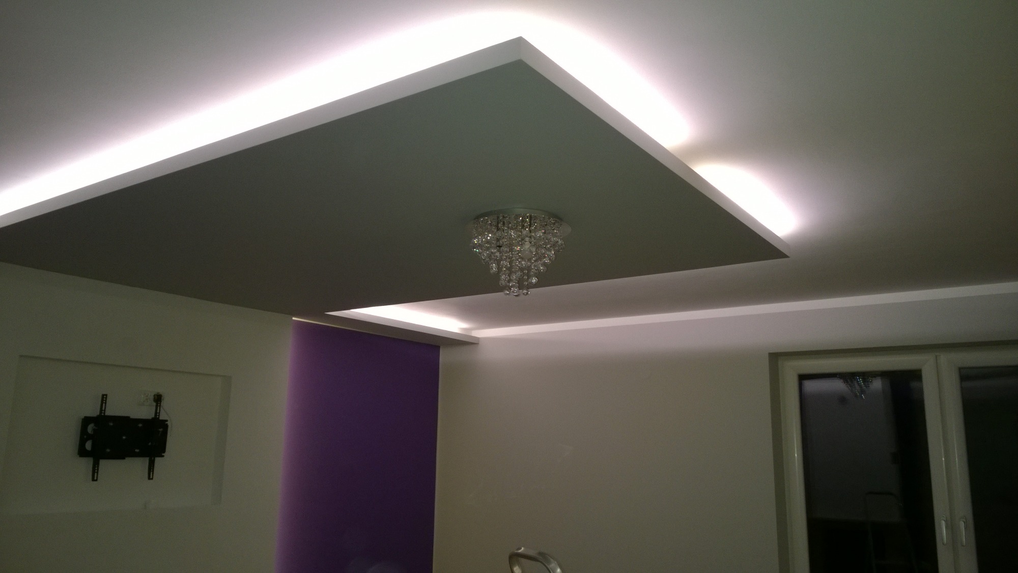 Sufit podwieszany z oświetleniem LED i kryształowym żyrandolem, ściana pomalowana na fioletowo, uchwyt na telewizor na ścianie.