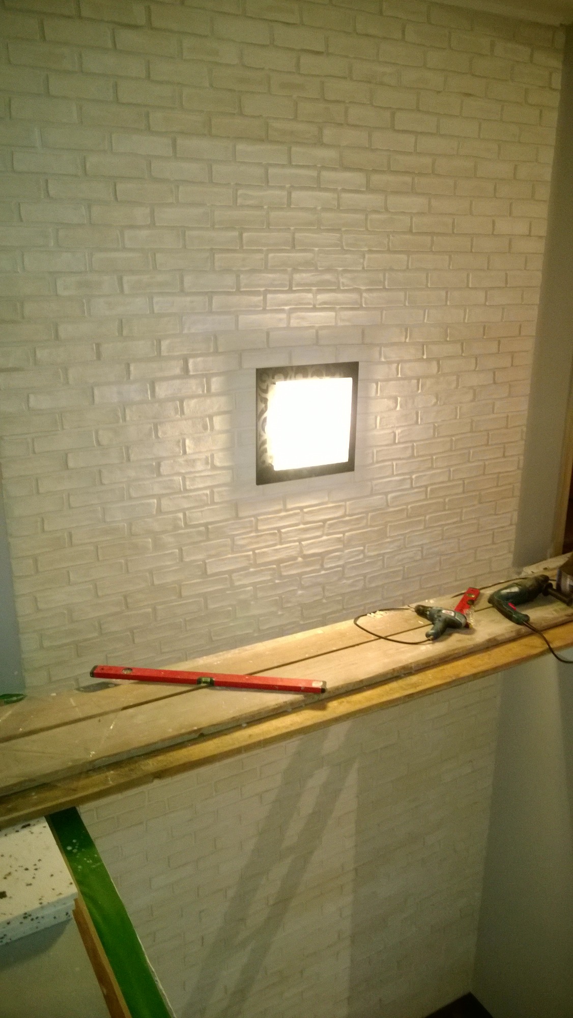 Ściana wyłożona białą cegłą z wbudowanym oświetleniem, na drewnianej półce poziomica i narzędzia elektryczne.