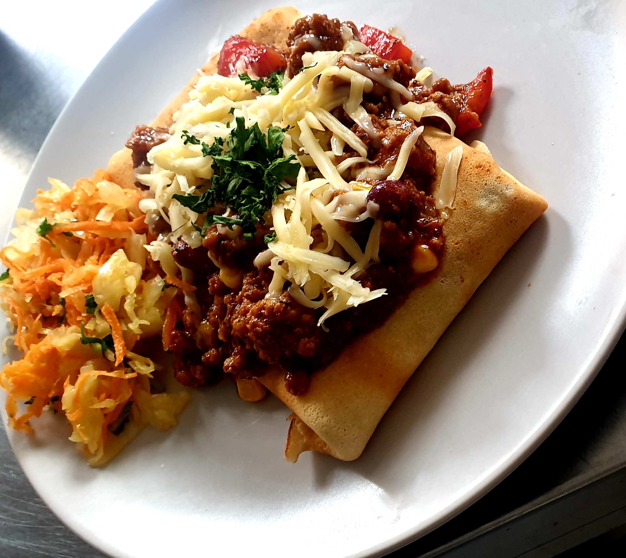 Naleśnik z mięsnym sosem chili, startym serem i posiekaną natką pietruszki, podany z surówką z marchwi i kapusty na białym talerzu.