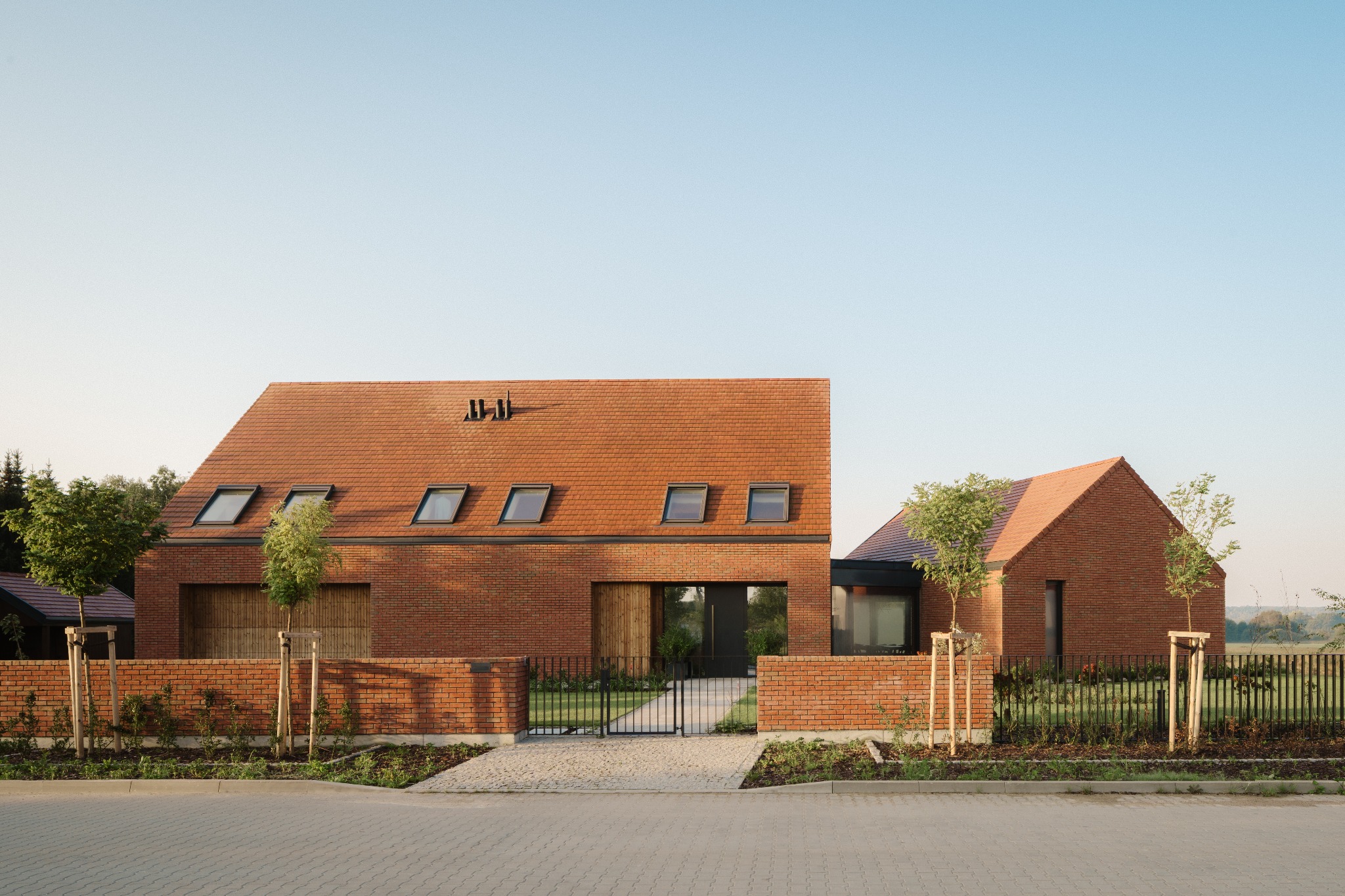 Nowoczesny dom z czerwonej cegły z dwuspadowym dachem i oknami dachowymi, otoczony niskim murem i zielenią. Minimalistyczna brama wejściowa. Ujęcie z poziomu ulicy.