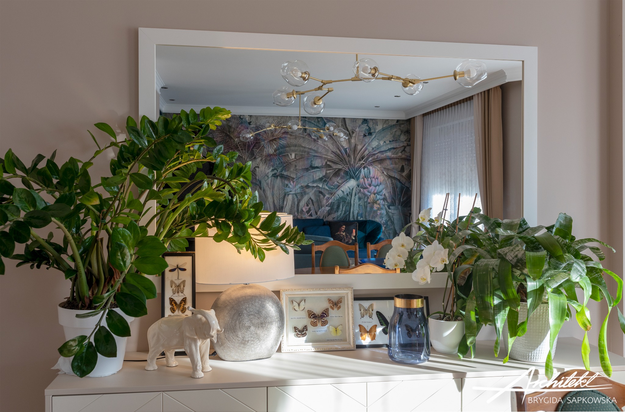 Pokój dzienny z tapetą w tropikalne wzory, widoczny w lustrze nad białą komodą. Na komodzie rośliny doniczkowe, biała figurka słonia, lampa ze srebrną podstawą oraz oprawione w ramki motyle. W tle...