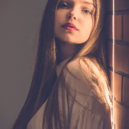 Portret młodej kobiety z długimi włosami, opierającej się o ceglaną ścianę w miękkim, bocznym świetle, z intensywnym spojrzeniem w obiektyw.