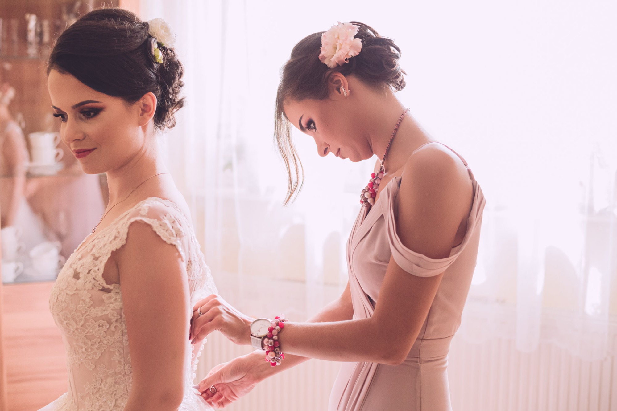 Panna młoda w koronkowej sukni ślubnej, z kwiatem we włosach, szykowana przez kobietę w pudrowej sukience, zapinającą guziki, Kraków.