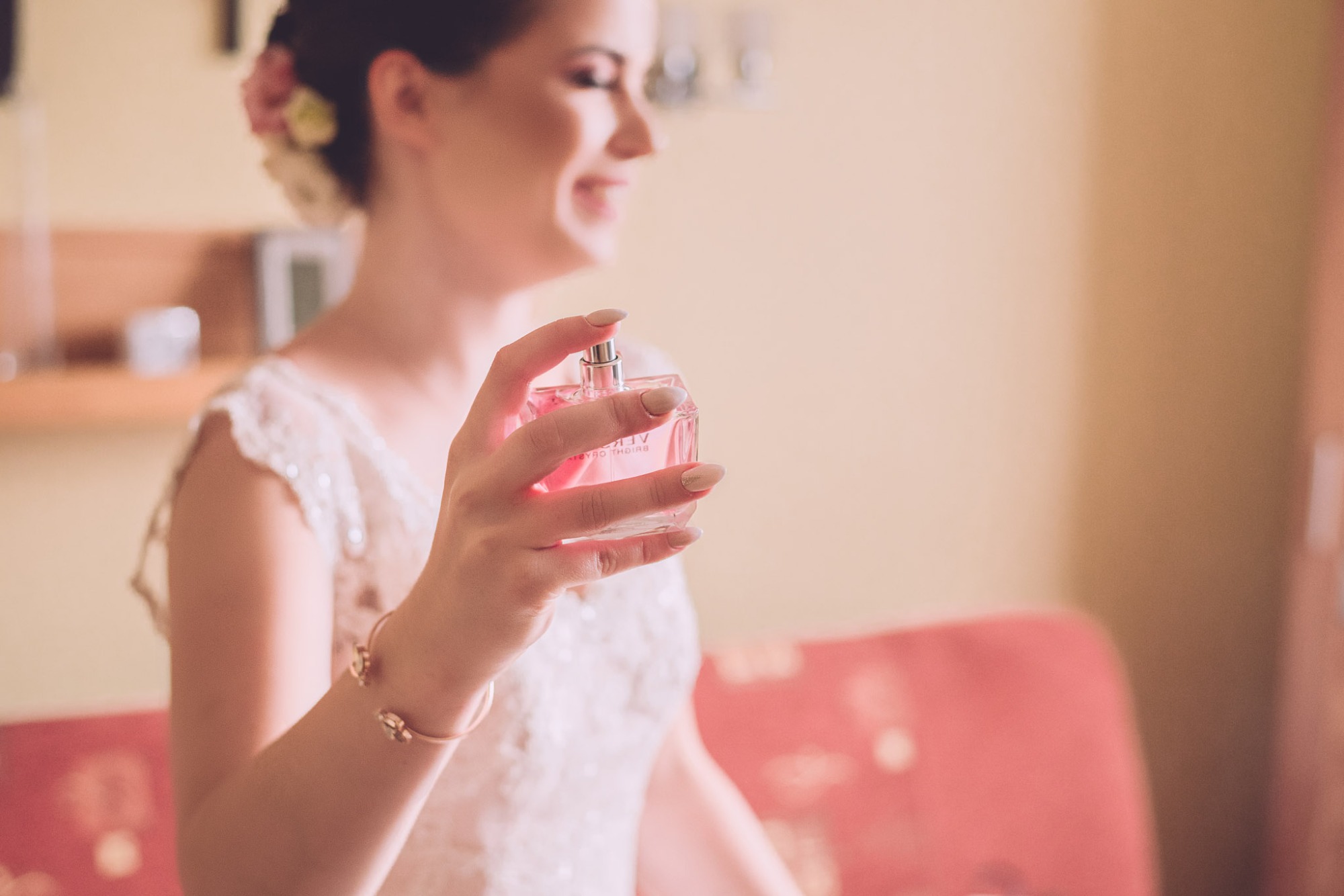 Uśmiechnięta panna młoda z kwiatami we włosach, w koronkowej sukni, trzymająca flakon perfum przed ślubem.