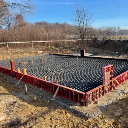 Zbrojenie płyty fundamentowej w szalunku systemowym, widoczne rury przepustowe, w tle drzewa i błękitne niebo.