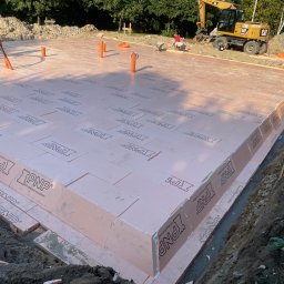 DM BUD Dariusz Marczykiewicz - Ocieplenie fundamentów budynku płytami XPS z widocznym wykopem, pracownikiem z odkurzaczem przemysłowym i koparką w tle.