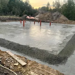 Świeżo wylana płyta fundamentowa z wystającymi rurami instalacyjnymi, widoczne deskowanie obrzeża i hałda ziemi w tle.