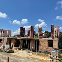 DM BUD Dariusz Marczykiewicz - Budowa domu z czerwonej cegły, widoczne szalunki na słupy, materiały budowlane składowane na placu budowy, błękitne niebo z chmurami w tle.