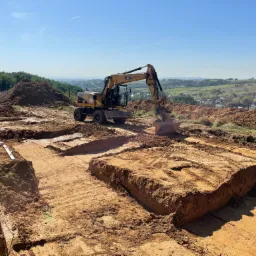 Żółta koparka CAT podczas prac ziemnych, przygotowująca teren pod fundamenty z widocznymi wykopami, poziomica i łata na skraju wykopu, w tle zielony krajobraz.