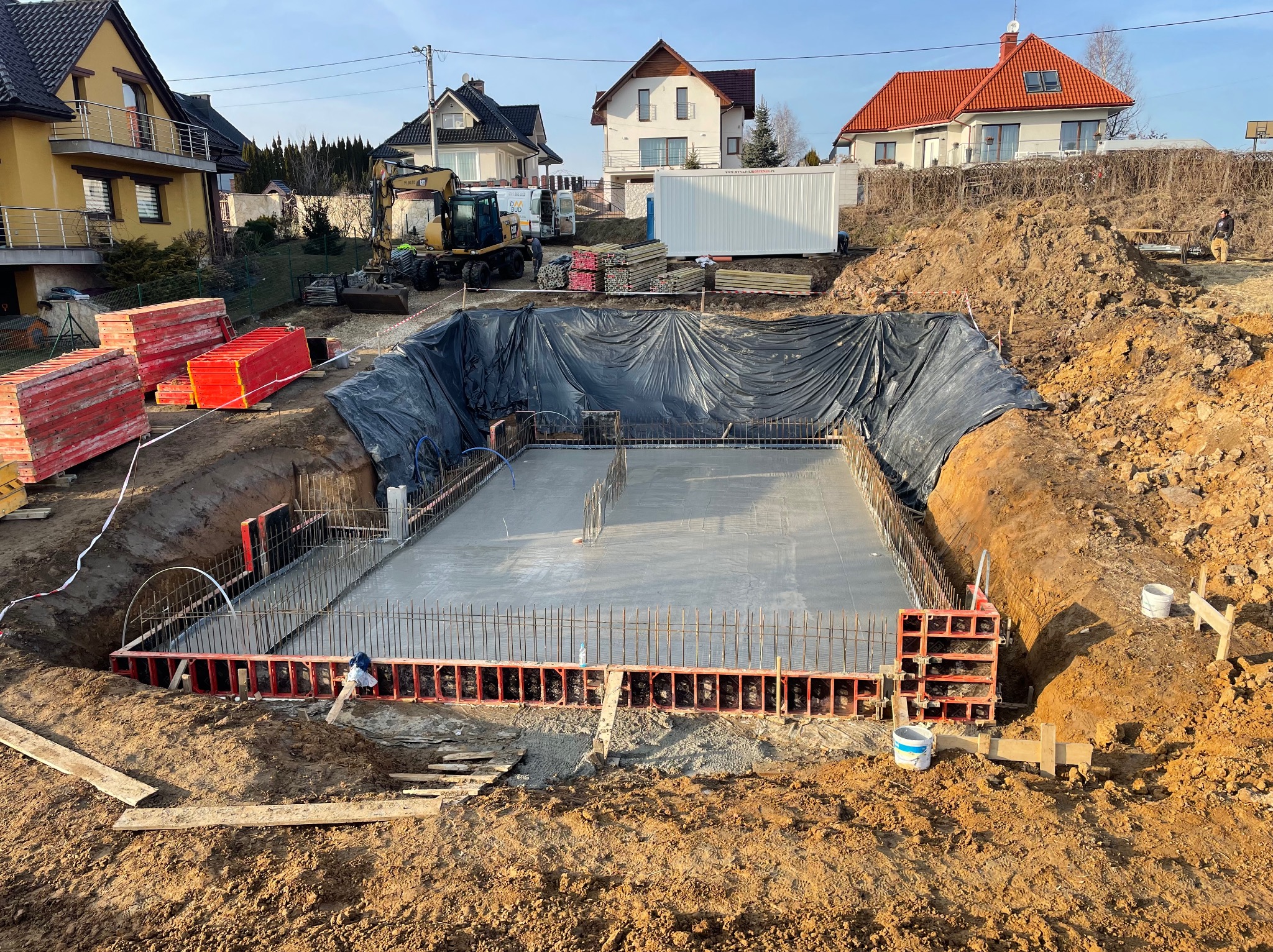 Wylany betonowy fundament zbrojony, otoczony szalunkiem w wykopie zabezpieczonym czarną folią, na tle domów i koparki.