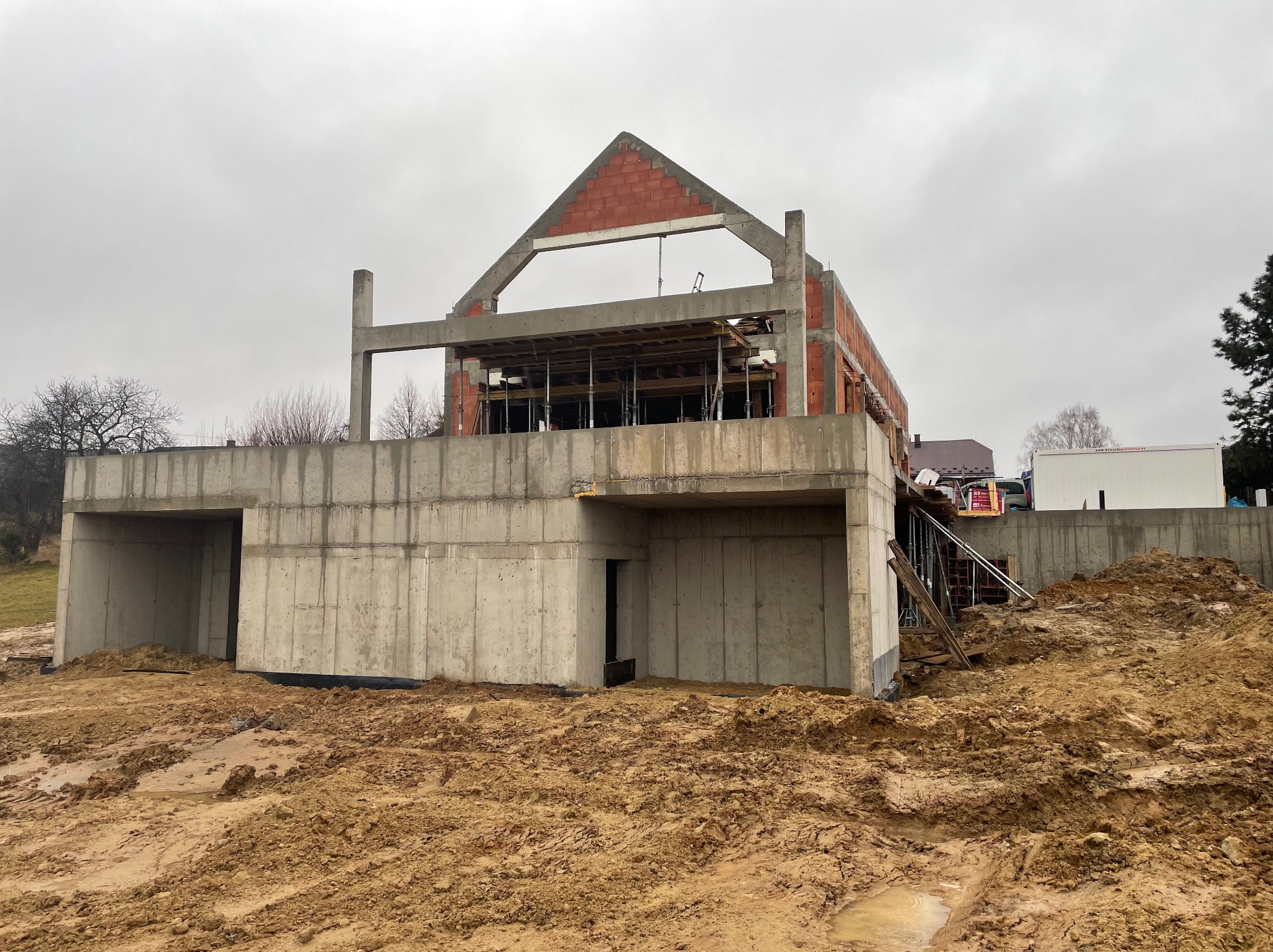 Surowy stan domu w trakcie budowy: widoczne betonowe fundamenty z otworami, ściany z czerwonej cegły wznoszące się do poziomu dachu, szalunki i podpory na górze, teren wokół rozkopany i błotnisty...