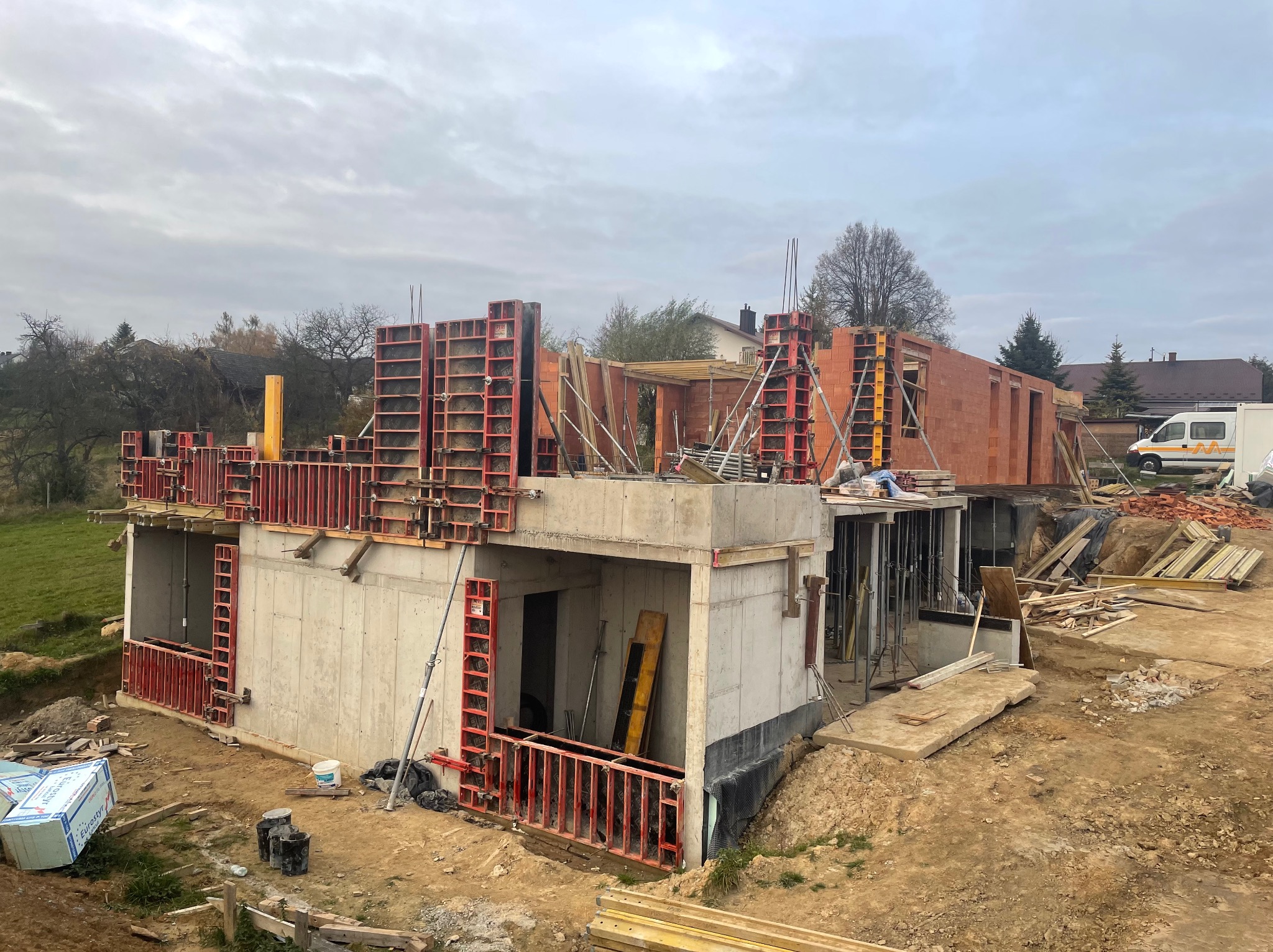 Budowa domu w trakcie realizacji: widoczne szalunki fundamentów, ściany z czerwonej cegły oraz materiały budowlane na placu budowy w pochmurny dzień.