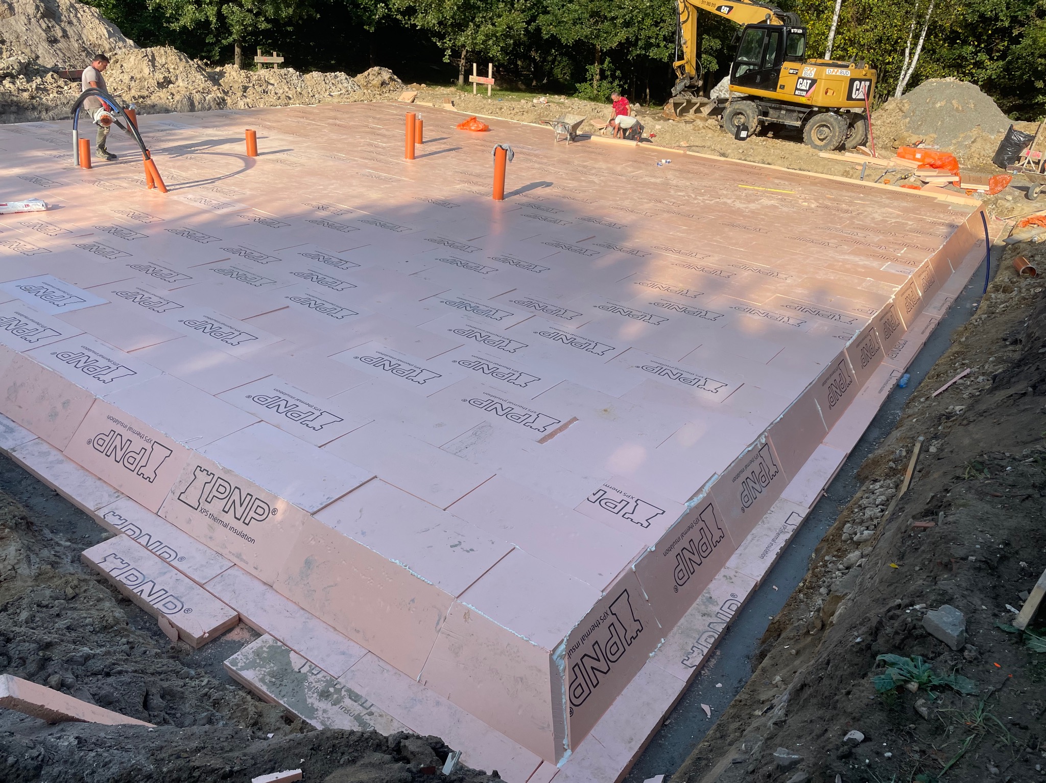 Ocieplenie fundamentów budynku płytami XPS z widocznym wykopem, pracownikiem z odkurzaczem przemysłowym i koparką w tle.