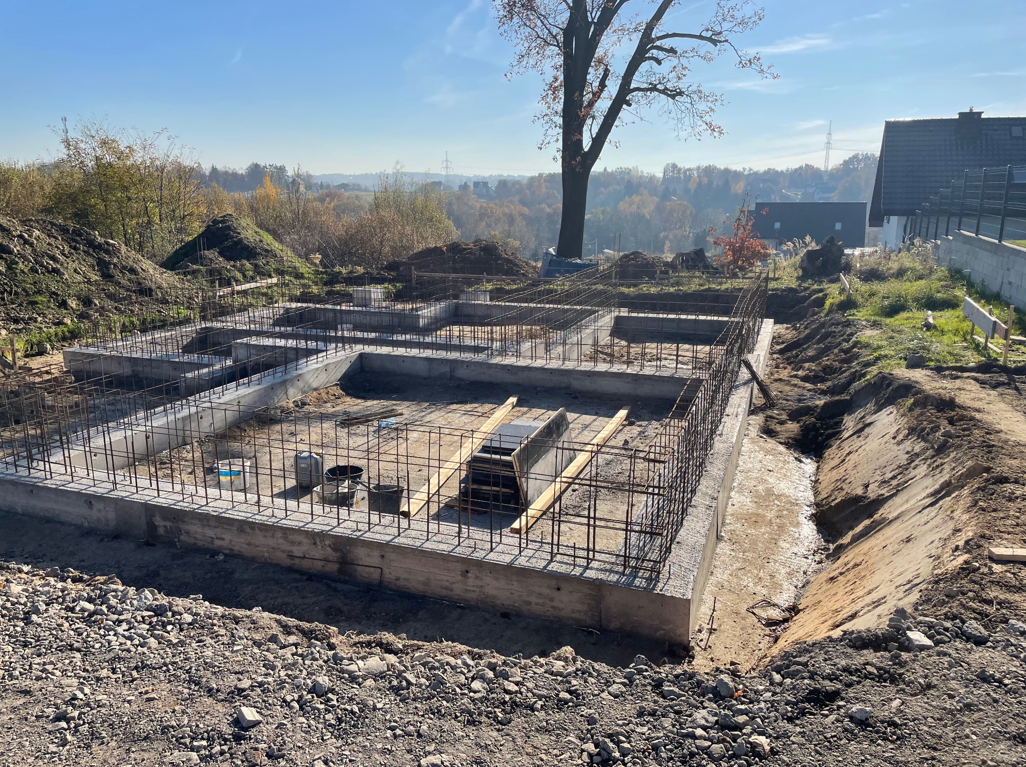 Zalane betonem fundamenty z wystającą siatką zbrojeniową na budowie domu jednorodzinnego, widoczne narzędzia i wykop pod fundamenty, w tle drzewa i zabudowa.