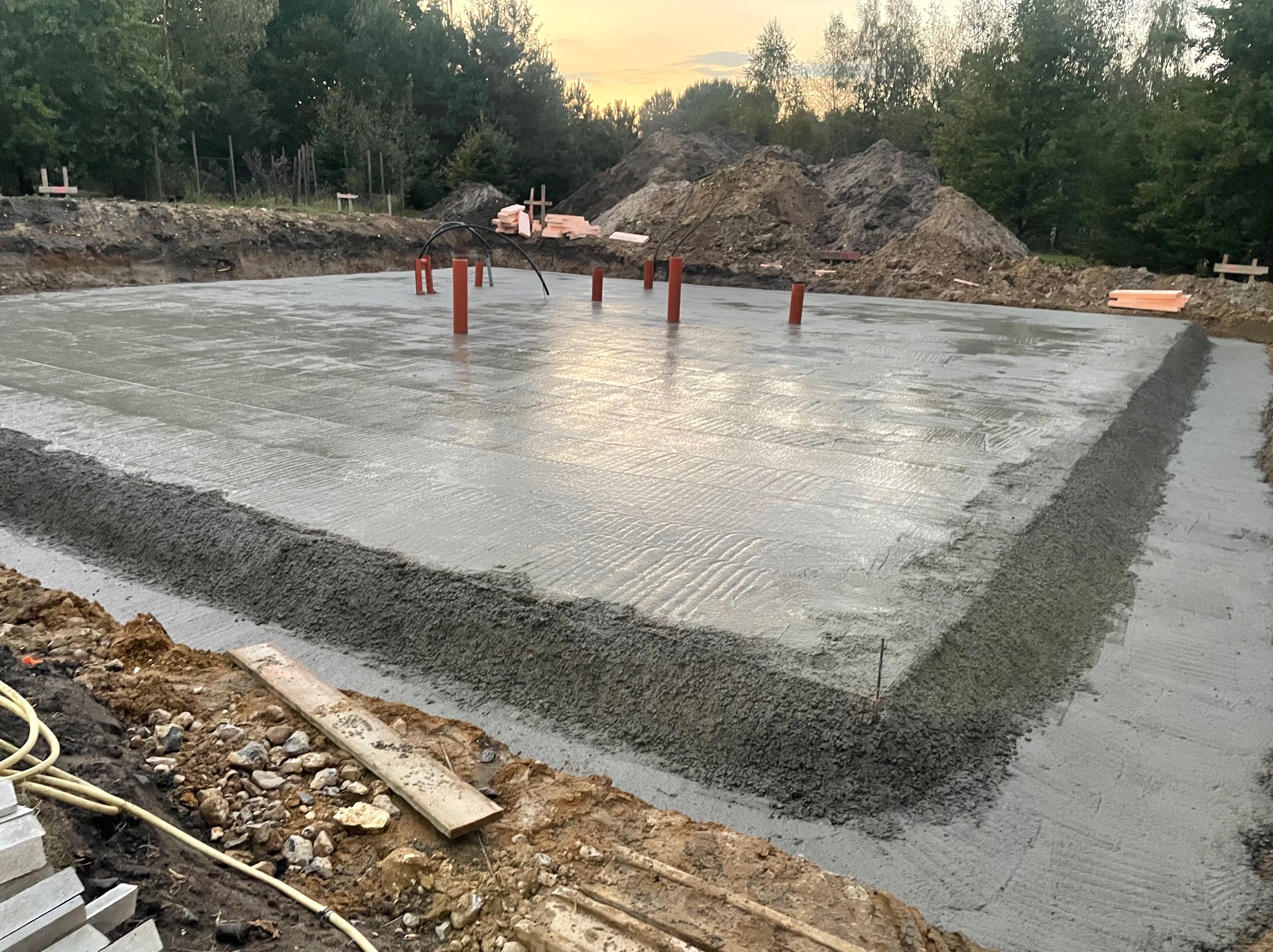 Świeżo wylana płyta fundamentowa z wystającymi rurami instalacyjnymi, widoczne deskowanie obrzeża i hałda ziemi w tle.