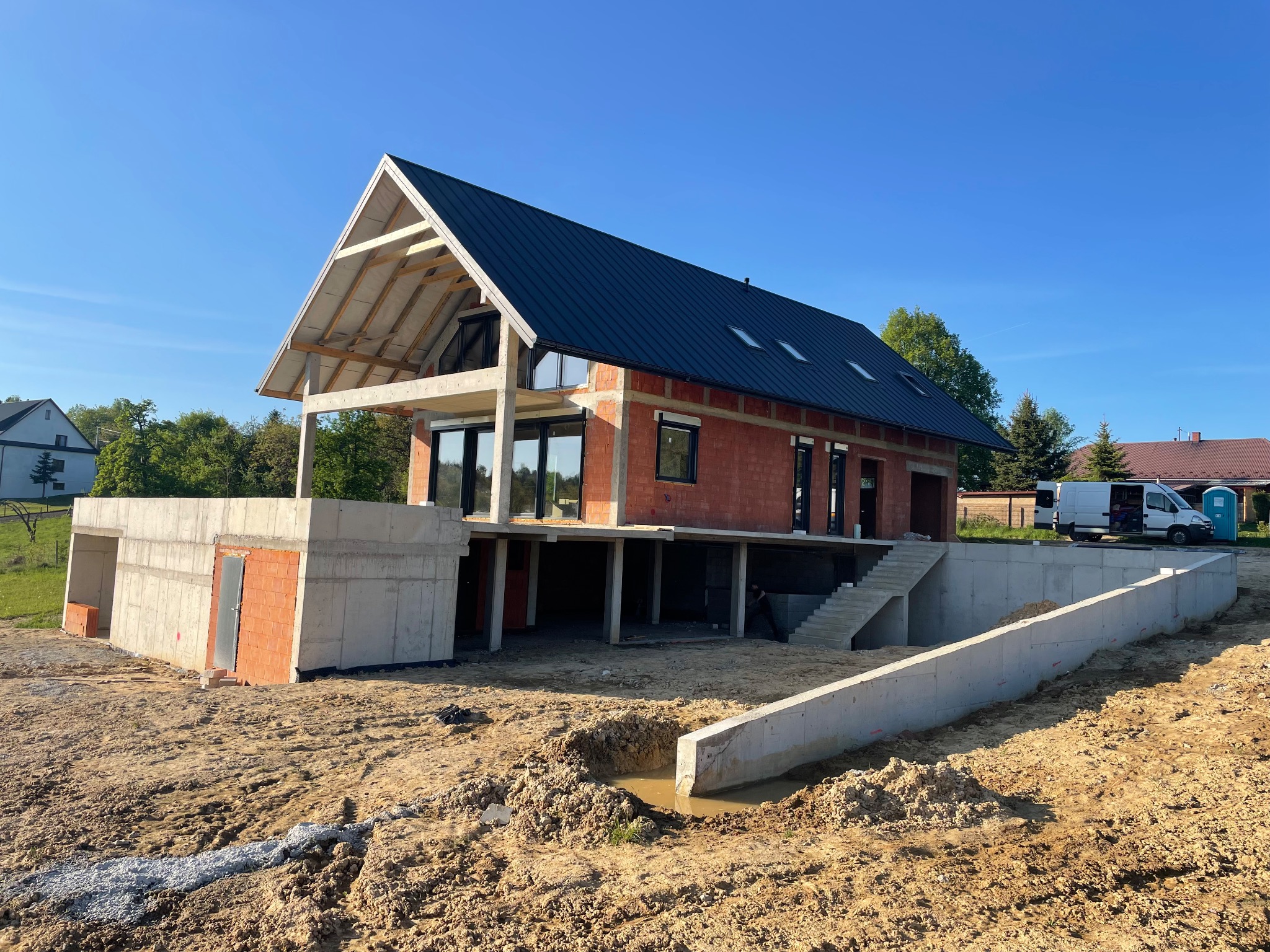 Surowy stan domu z dachem, widoczne bloczki i betonowe fundamenty. Teren wokół domu w trakcie prac ziemnych, częściowo wykopany. Błękitne niebo w tle.