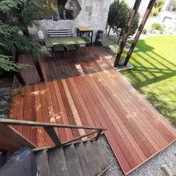 Widok z góry na taras z desek kompozytowych w odcieniach brązu, połączony ze schodami i otoczony zielenią trawnika i drzew, z widocznym zestawem mebli ogrodowych.