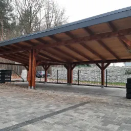 Zadaszenie drewniane z ciemną, metalową obróbką, wsparte na solidnych, brązowych słupach, na wybrukowanym podłożu z widocznymi oponami i fragmentem ogrodzenia.