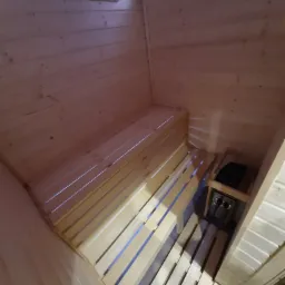 Wnętrze sauny wykonane z jasnego drewna, z ławkami ułożonymi wzdłuż ścian i piecem elektrycznym w rogu.