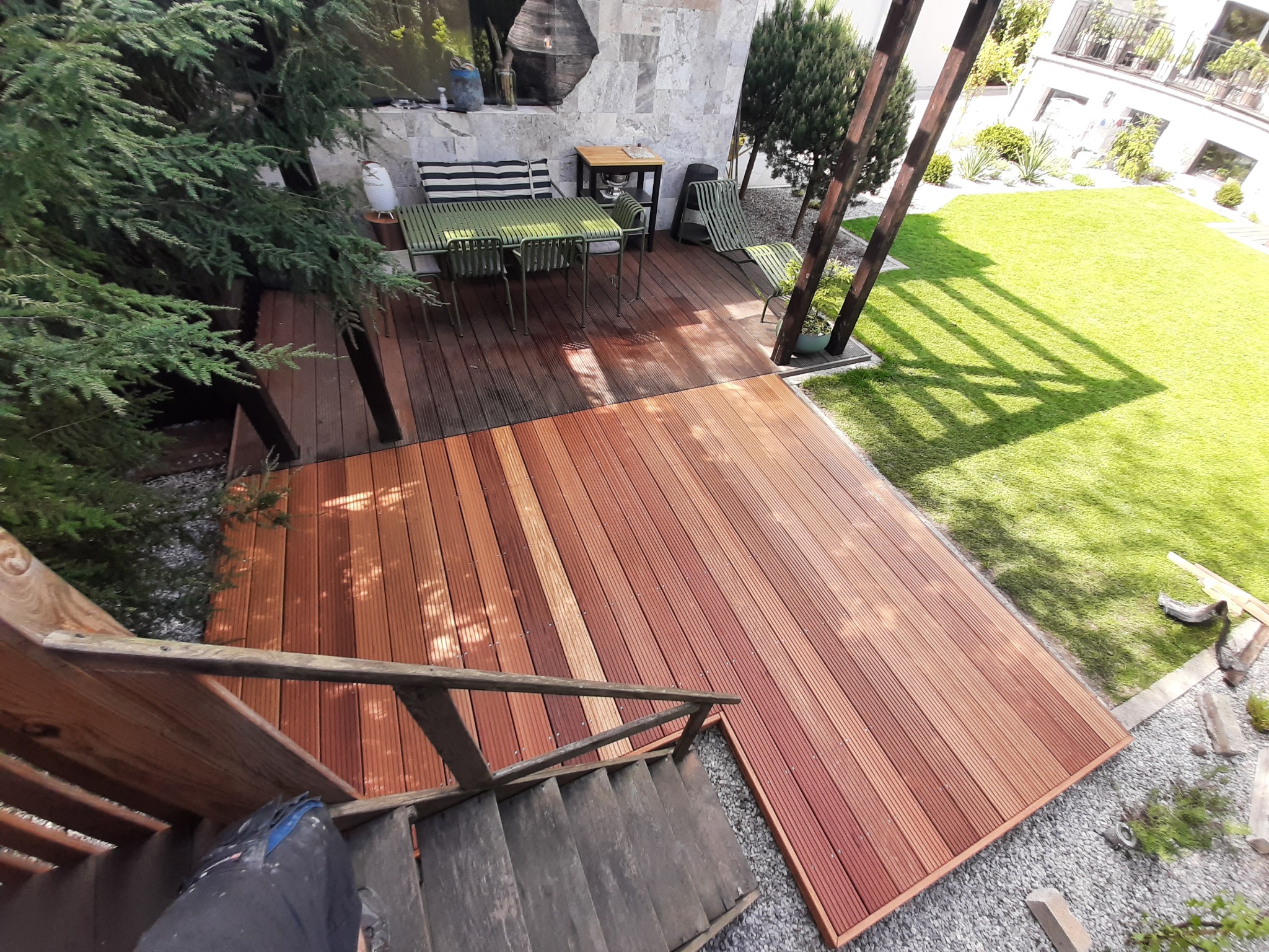 Widok z góry na taras z desek kompozytowych w odcieniach brązu, połączony ze schodami i otoczony zielenią trawnika i drzew, z widocznym zestawem mebli ogrodowych.