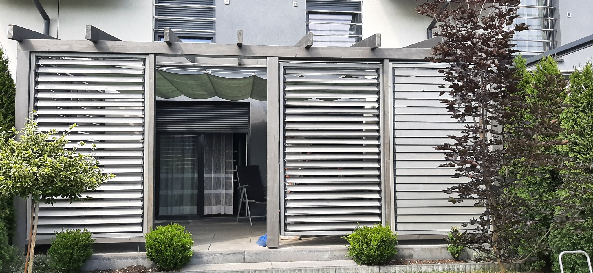 Nowoczesna, szara konstrukcja z regulowanymi lamelami, tworząca zacienione przejście do wnętrza, otoczona zielenią krzewów i drzew, z widocznym fragmentem domu w tle.