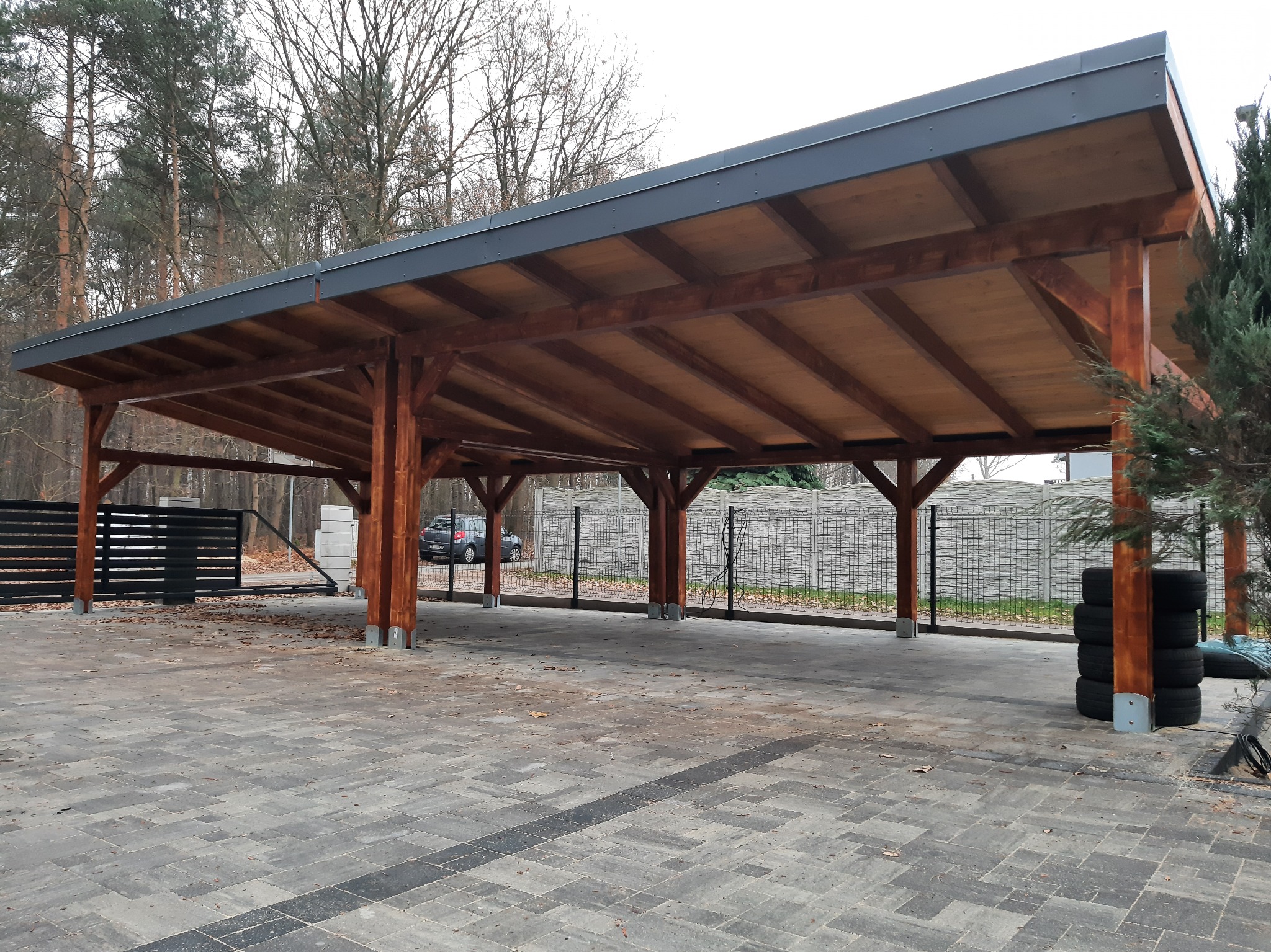 Zadaszenie drewniane z ciemną, metalową obróbką, wsparte na solidnych, brązowych słupach, na wybrukowanym podłożu z widocznymi oponami i fragmentem ogrodzenia.