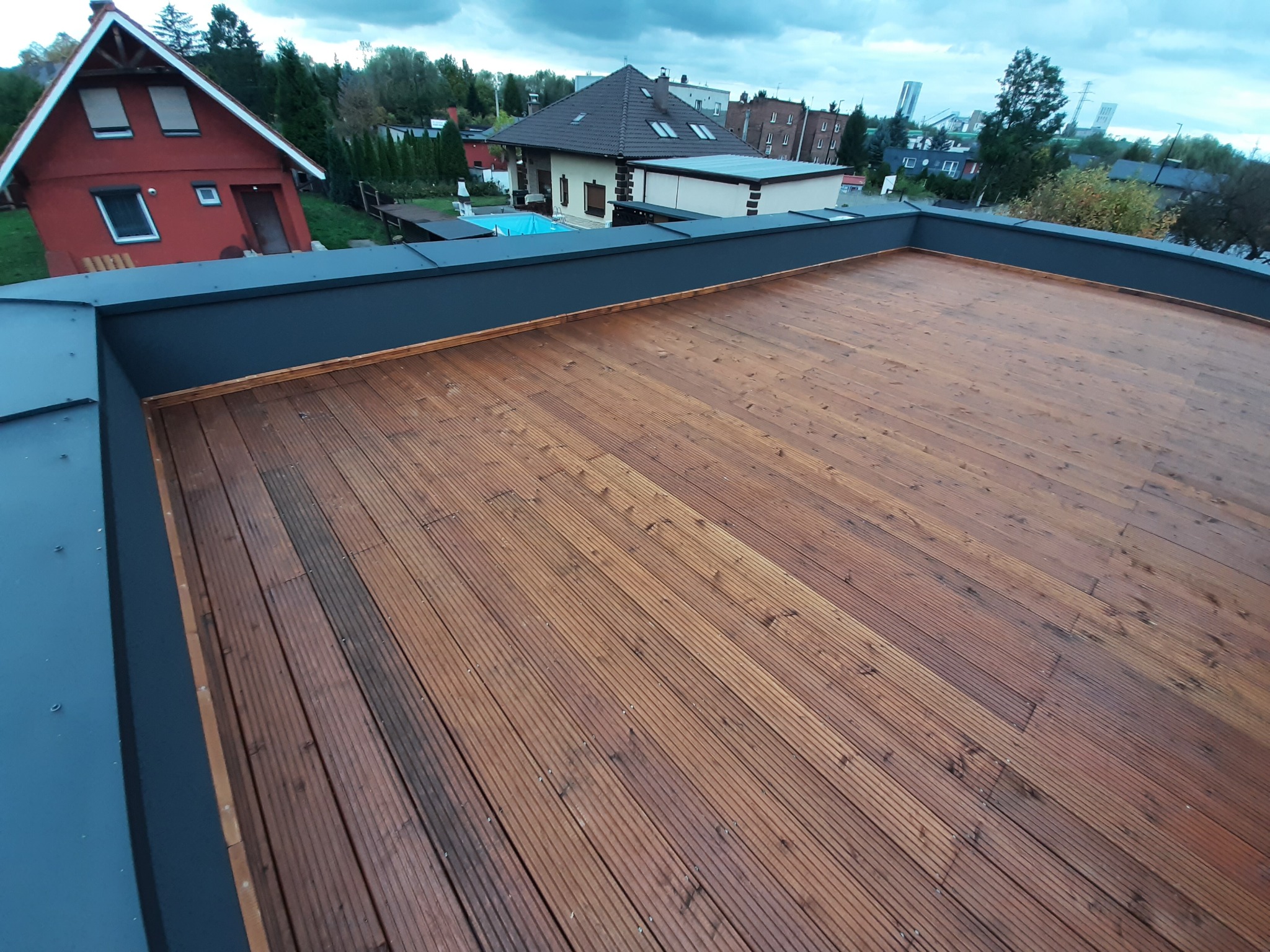 Nowoczesny brązowy taras na dachu z widokiem na domy, basen i zabudowania przemysłowe w tle.