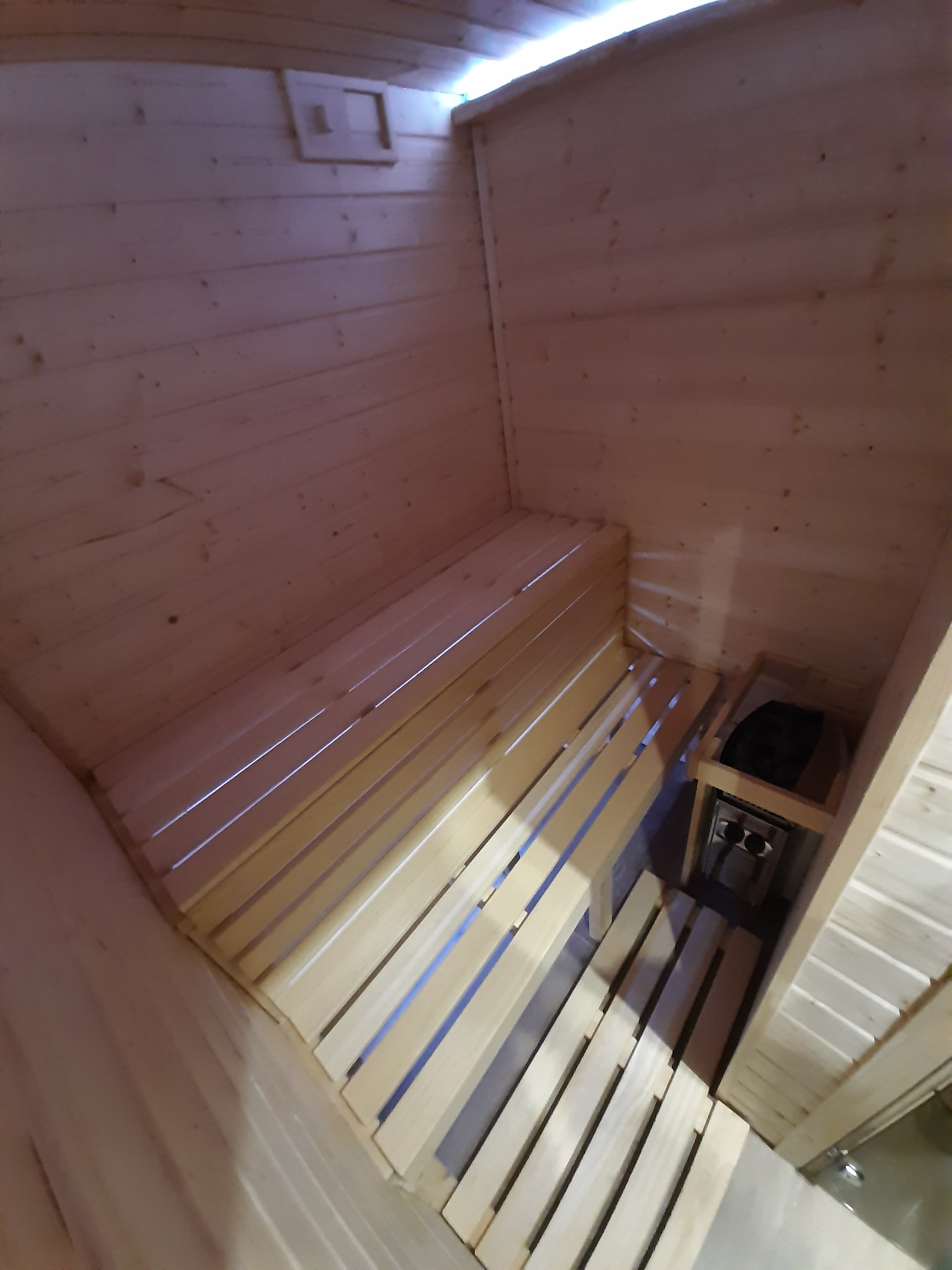 Wnętrze sauny wykonane z jasnego drewna, z ławkami ułożonymi wzdłuż ścian i piecem elektrycznym w rogu.