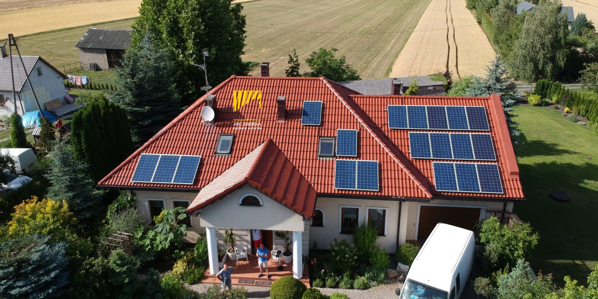 Dom jednorodzinny z czerwoną dachówką i panelami fotowoltaicznymi, widok z lotu ptaka, na dachu widoczne logo firmy, w tle pole uprawne i zabudowania gospodarcze.