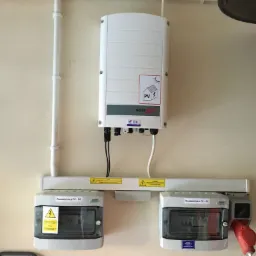 Biały inwerter fotowoltaiczny z rozdzielnicami PV-DC i PV-AC zamontowany na beżowej ścianie, widoczne przewody i oznaczenia ostrzegawcze.