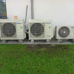 Trzy jednostki zewnętrzne klimatyzacji Panasonic zamontowane na betonowych podstawach z wysypanym dookoła kamieniem, na tle trawnika i ściany budynku.