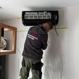 Klima Siedlce Tomasz Pawlik - Instalator na drabinie montuje czarny klimatyzator na ścianie, zabezpieczonej folią ochronną. Widoczna część obudowy i filtry urządzenia.