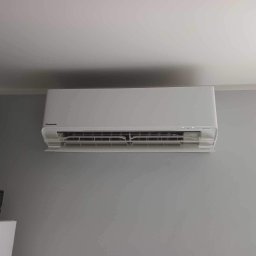 Klima Siedlce Tomasz Pawlik - Biała klimatyzacja Panasonic zamontowana na szarej ścianie w pomieszczeniu, widoczne fragmenty sufitu i podłogi, minimalistyczny design.