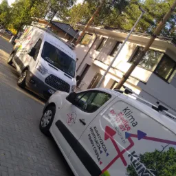 Dwa białe busy firmy Klima Siedlce z reklamami klimatyzacji, wentylacji i pomp ciepła zaparkowane na tle budynku mieszkalnego w słoneczny dzień.