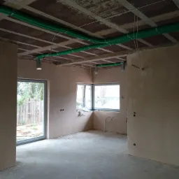 Wnętrze w stanie surowym z widoczną instalacją wentylacyjną w postaci zielonych rur pod sufitem, punktowe oświetlenie zamontowane do stropu, okna z widokiem na zewnątrz i przygotowane punkty...