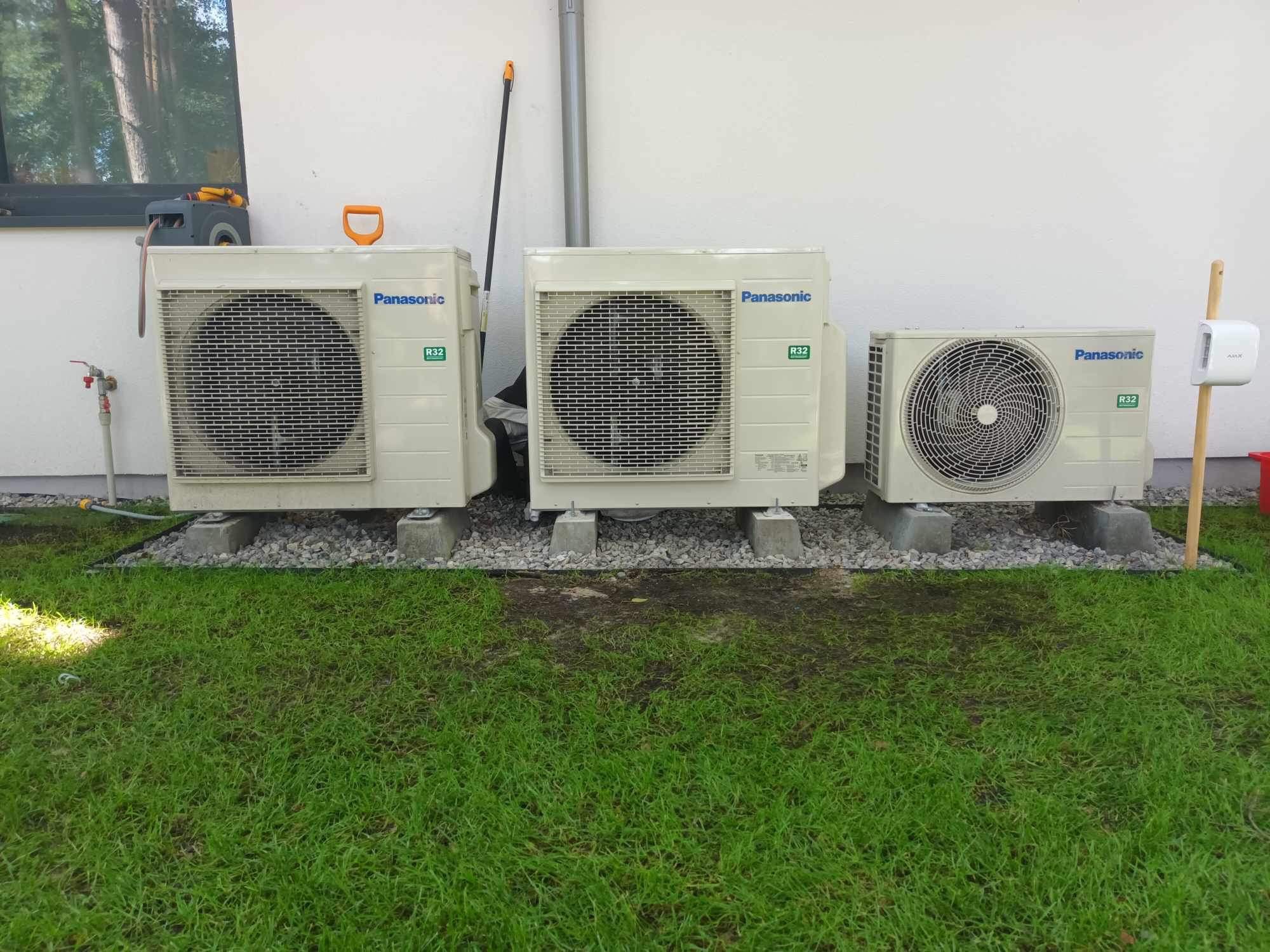 Trzy jednostki zewnętrzne klimatyzacji Panasonic zamontowane na betonowych podstawach z wysypanym dookoła kamieniem, na tle trawnika i ściany budynku.