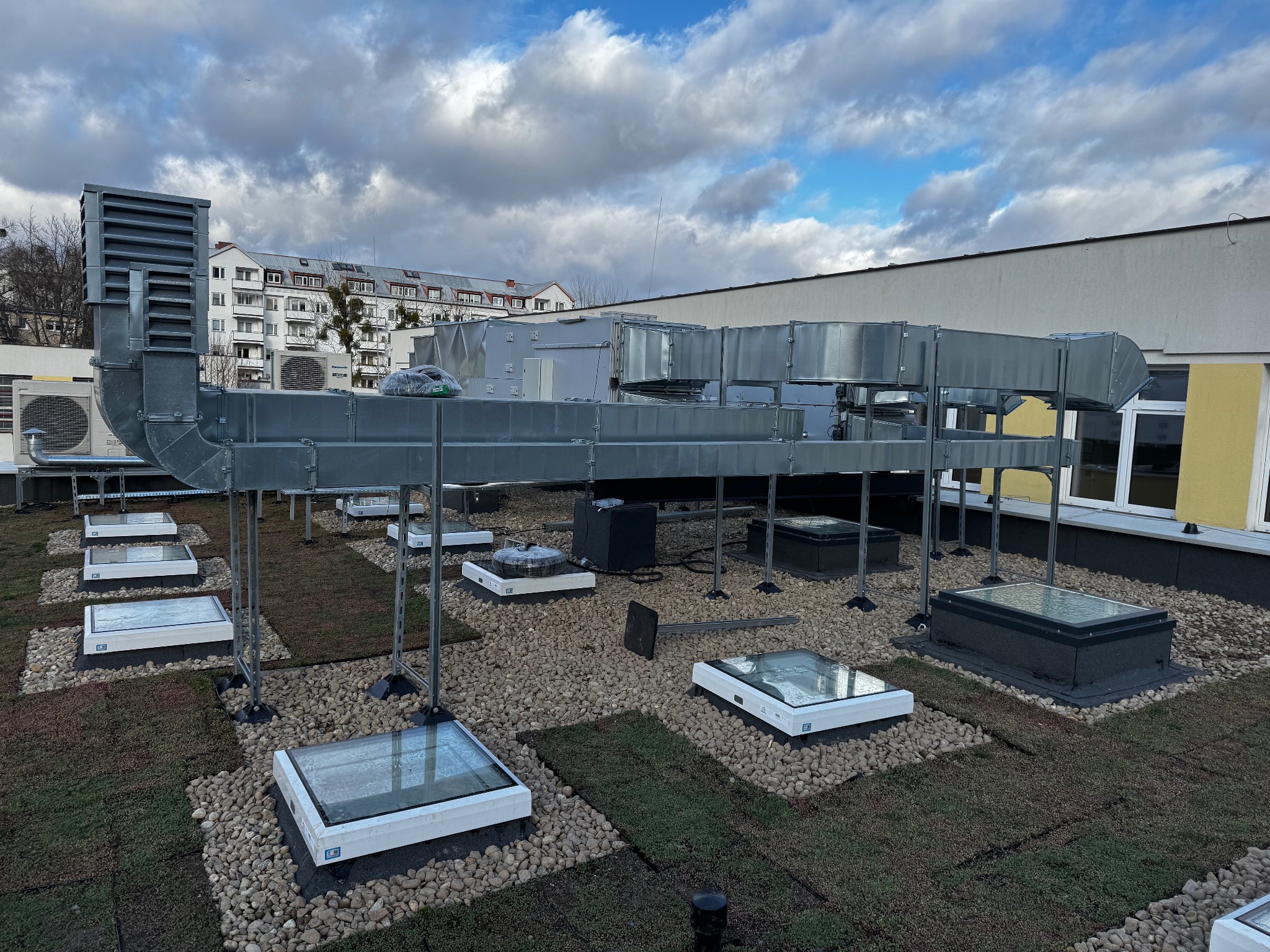 Szare kanały wentylacyjne na zielonym dachu budynku z oknami dachowymi; widok z góry.