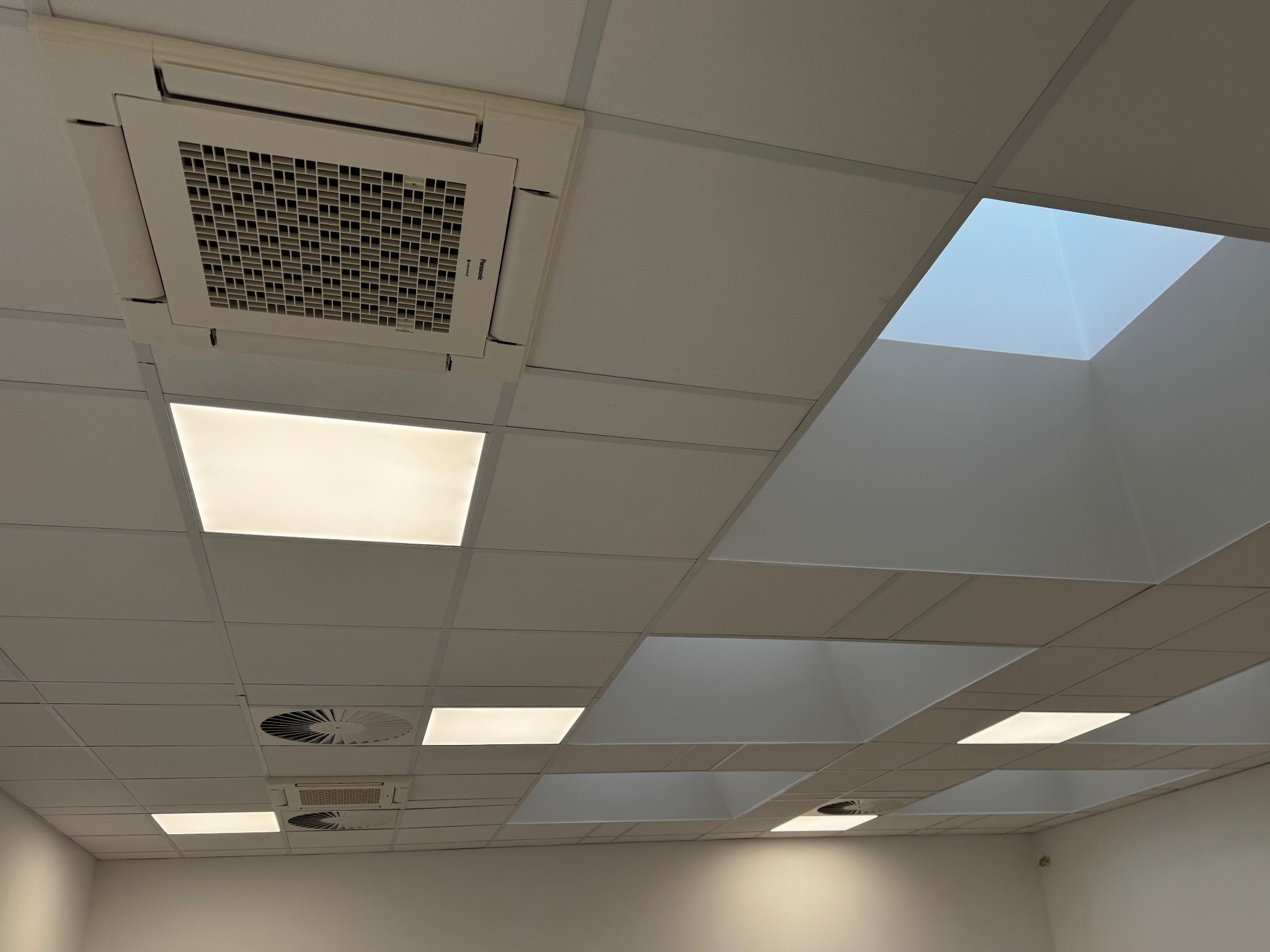 Sufit kasetonowy z wbudowaną klimatyzacją kasetonową, oświetleniem LED i elementami wentylacji. Widok z dołu, oświetlenie mieszane.
