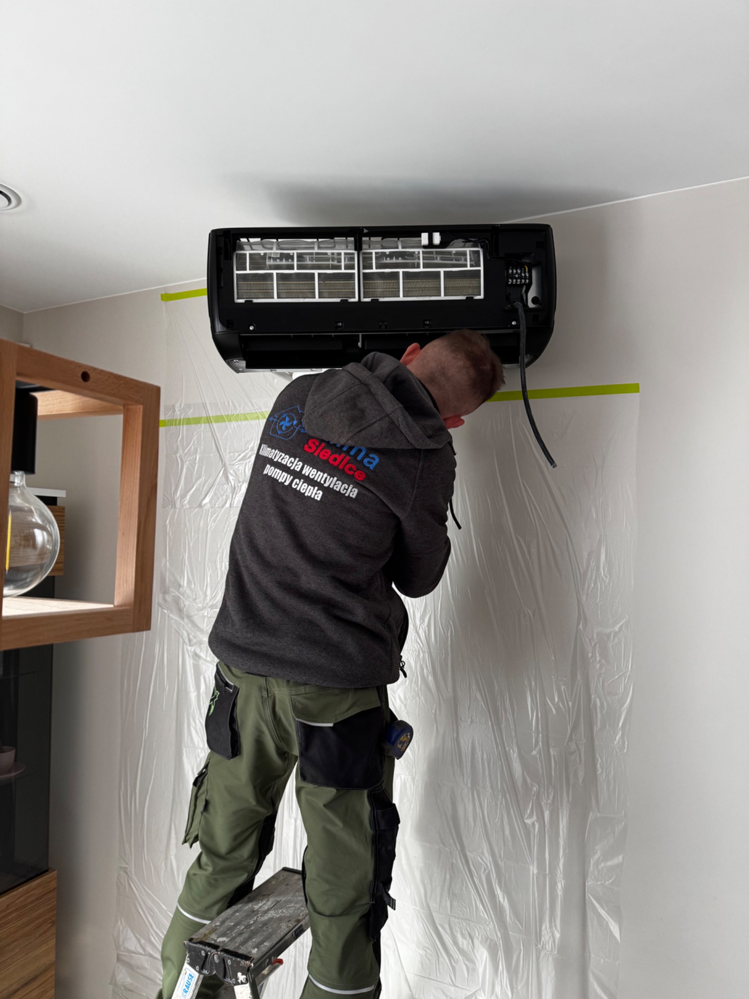 Instalator na drabinie montuje czarny klimatyzator na ścianie, zabezpieczonej folią ochronną. Widoczna część obudowy i filtry urządzenia.