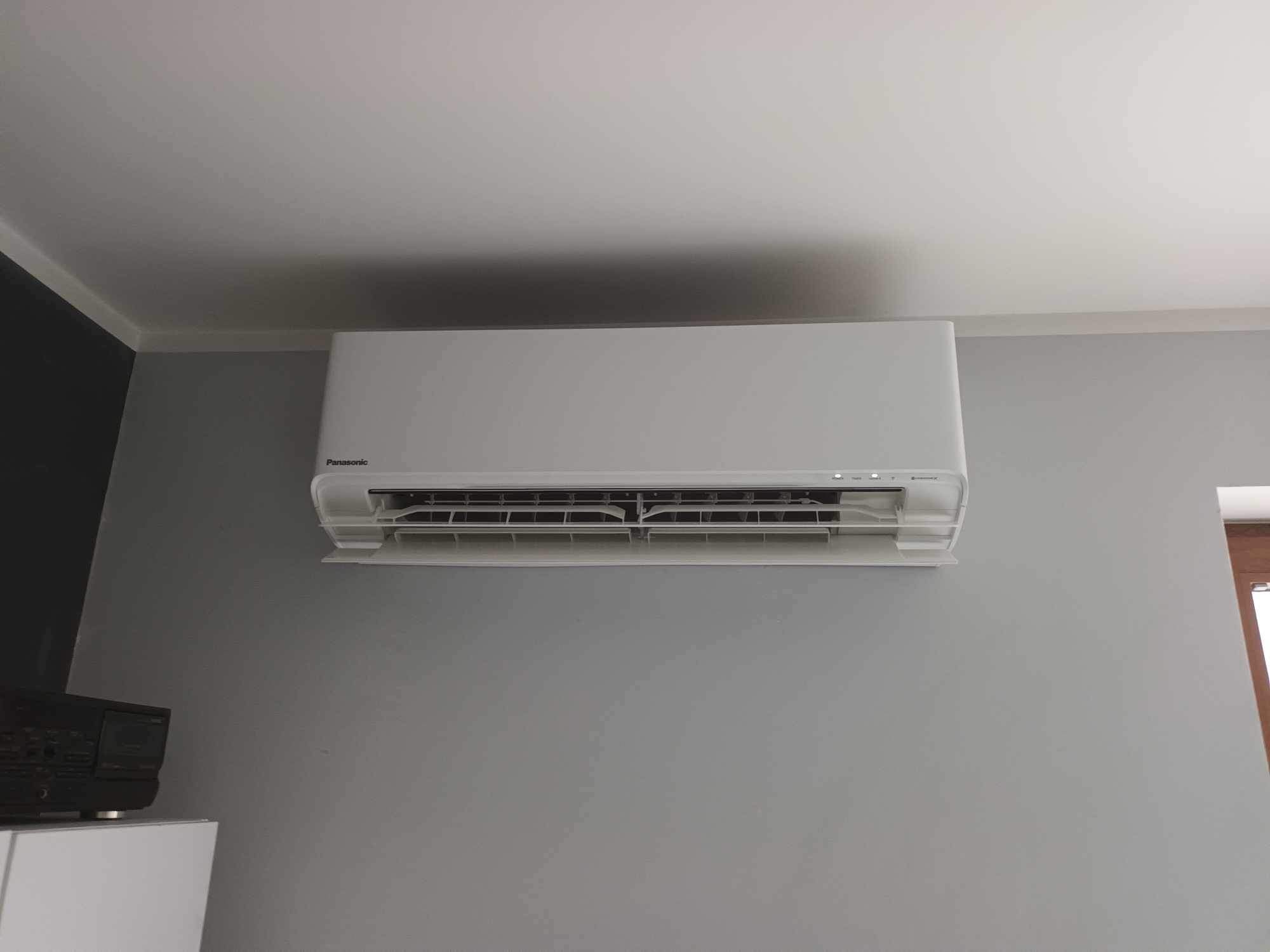 Biała klimatyzacja Panasonic zamontowana na szarej ścianie w pomieszczeniu, widoczne fragmenty sufitu i podłogi, minimalistyczny design.