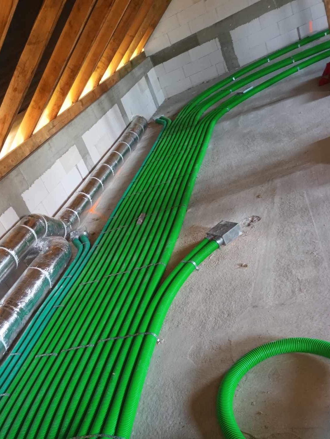 Zielone, karbowane rury wentylacyjne ułożone równolegle na betonowej podłodze poddasza. Rury z folią aluminiową po lewej stronie. Widoczne drewniane elementy konstrukcji dachu.
