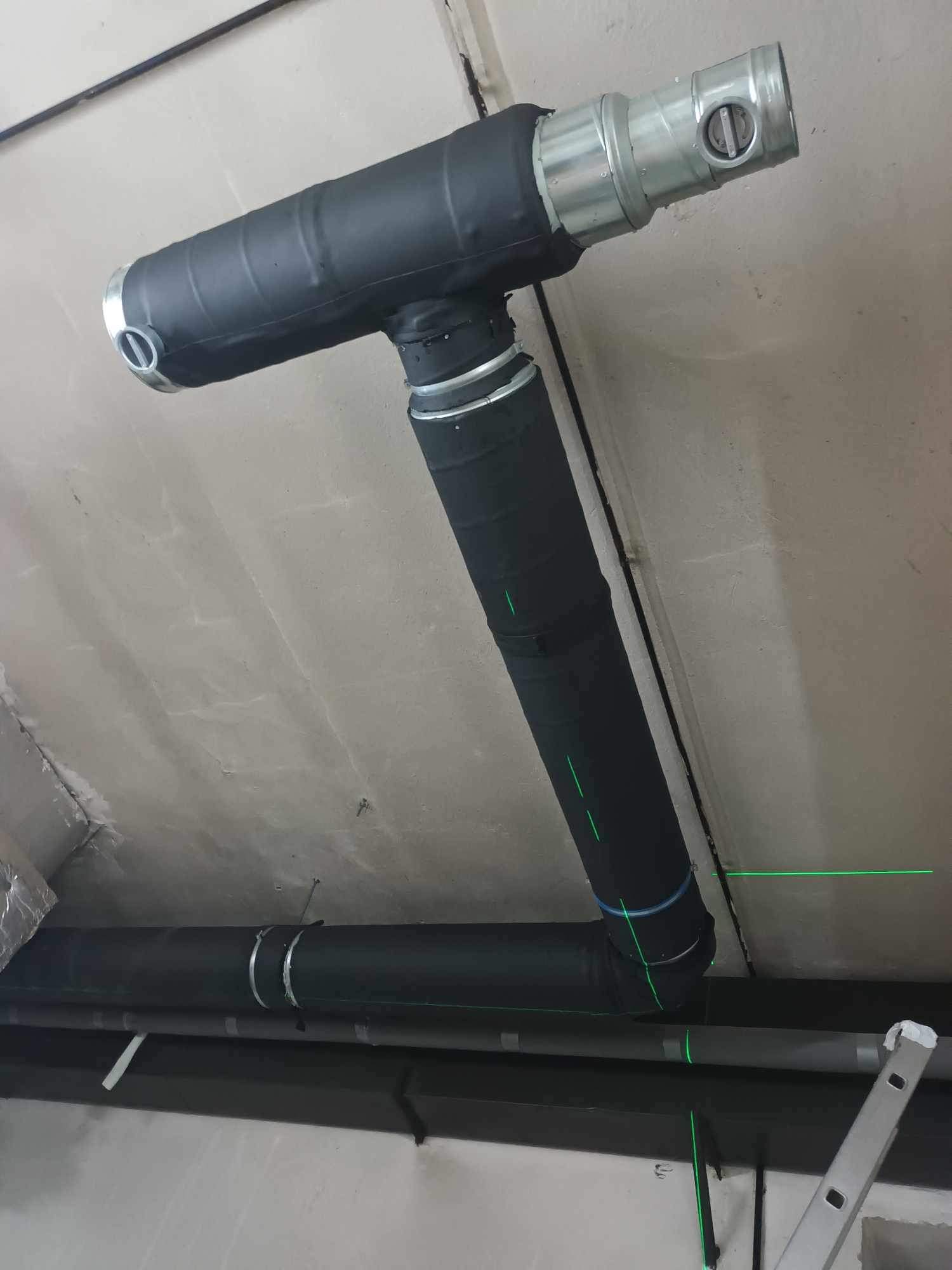 Czarna, zaizolowana rura wentylacyjna pod sufitem, z widocznymi metalowymi elementami i zielonym laserem poziomującym. Fragment drabiny w prawym dolnym rogu.