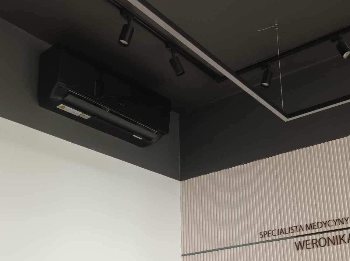 Czarny klimatyzator ścienny na tle białej ściany i czarnego sufitu z oświetleniem punktowym. Widoczny fragment ściany z napisem 'Specjalista medycyny'.