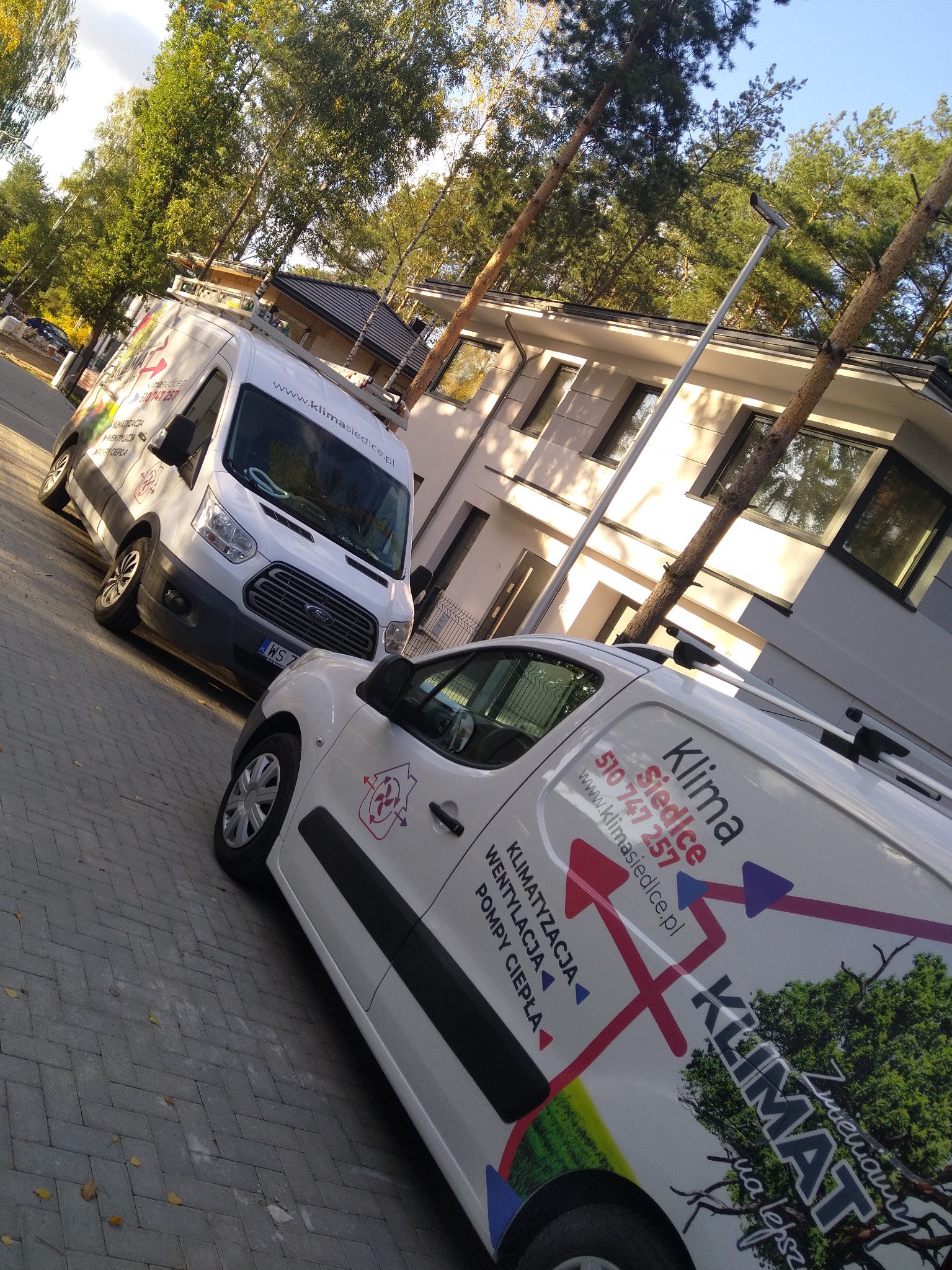 Dwa białe busy firmy Klima Siedlce z reklamami klimatyzacji, wentylacji i pomp ciepła zaparkowane na tle budynku mieszkalnego w słoneczny dzień.