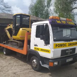Żółta mini koparka Komatsu PC35 na pomarańczowej platformie lawety pomocy drogowej MAN, zaparkowanej na piaszczystym terenie, z widocznym numerem telefonu kontaktowego na kabinie.