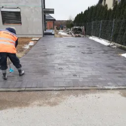 Układanie szarej kostki brukowej na podjeździe do domu jednorodzinnego, pracownik w pomarańczowej kamizelce zamiata kostkę, widoczne studzienki kanalizacyjne.