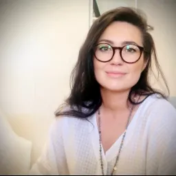 Portret kobiety w okularach, uśmiechniętej, w jasnej bluzce, siedzącej na fotelu w gabinecie.